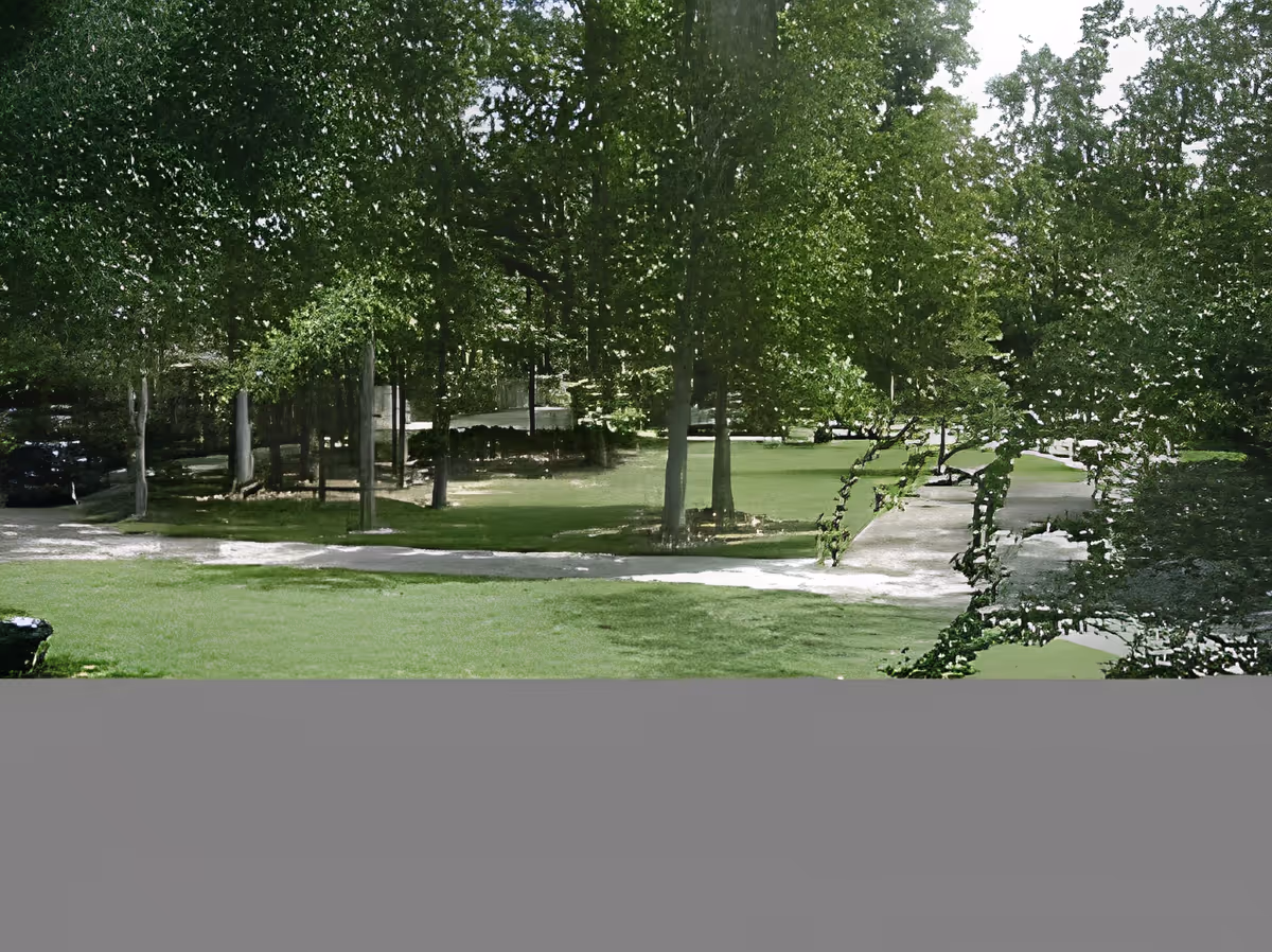 A green outdoor area with several tall trees and a paved pathway winding through the grass. The scene is bright with sunlight filtering through the leaves, creating a peaceful and natural environment.