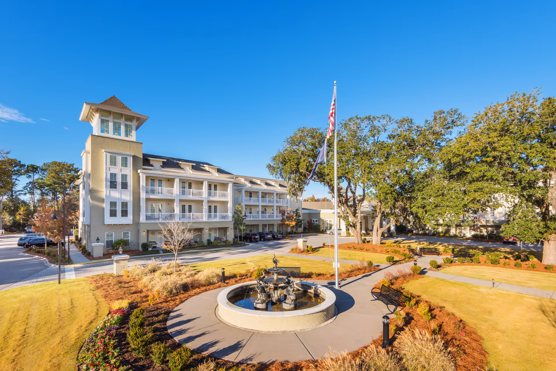 Exterior view of South Bay At Mount Pleasant facility featuring a multi-story building with balconies, a central fountain surrounded by a circular walkway, benches, landscaped gardens, and tall trees under a clear blue sky.