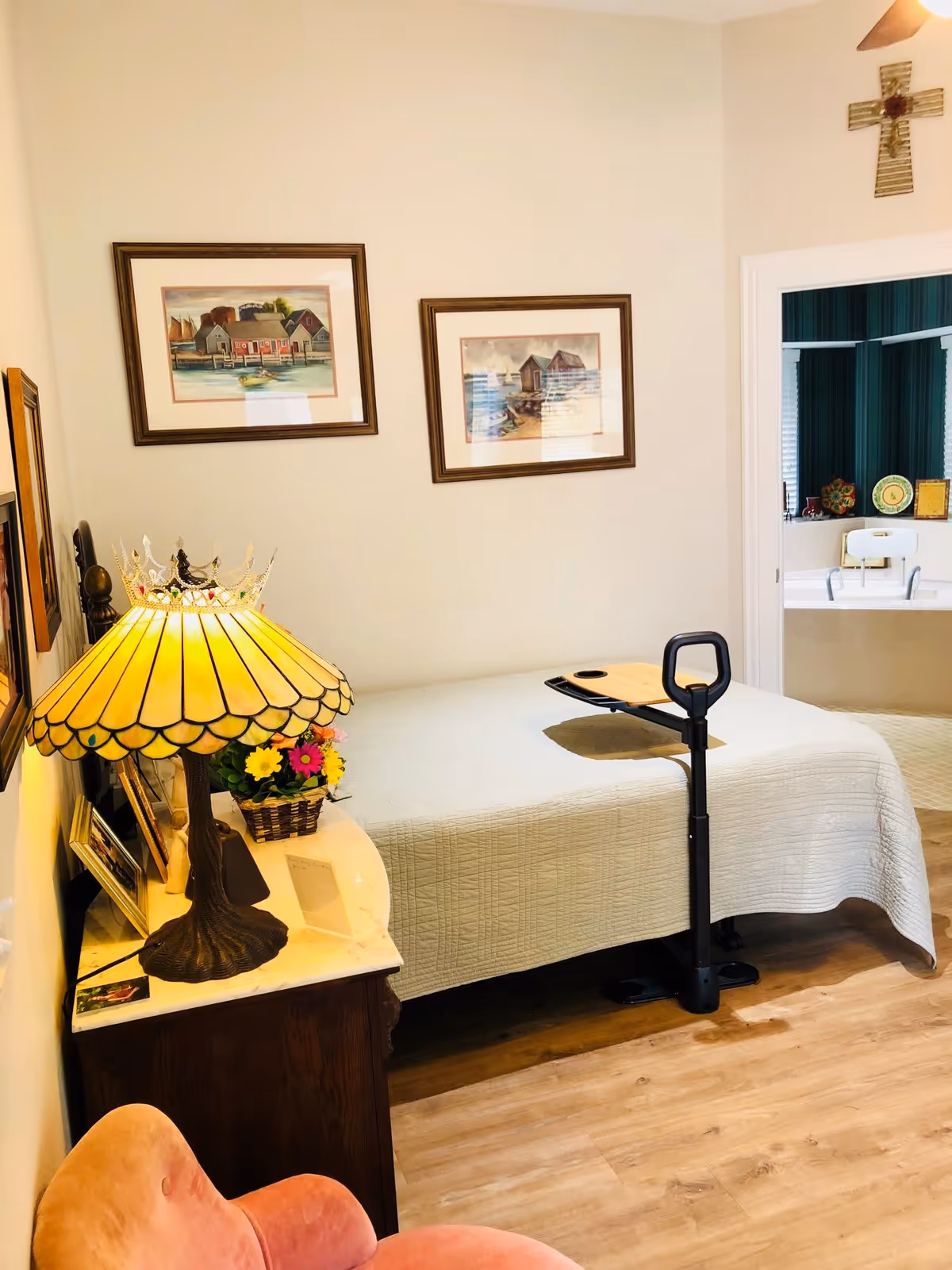 Small bedroom with a single bed, bedside table holding a stained-glass lamp and flowers, an overbed tray, and a view into an adjacent bathroom.
