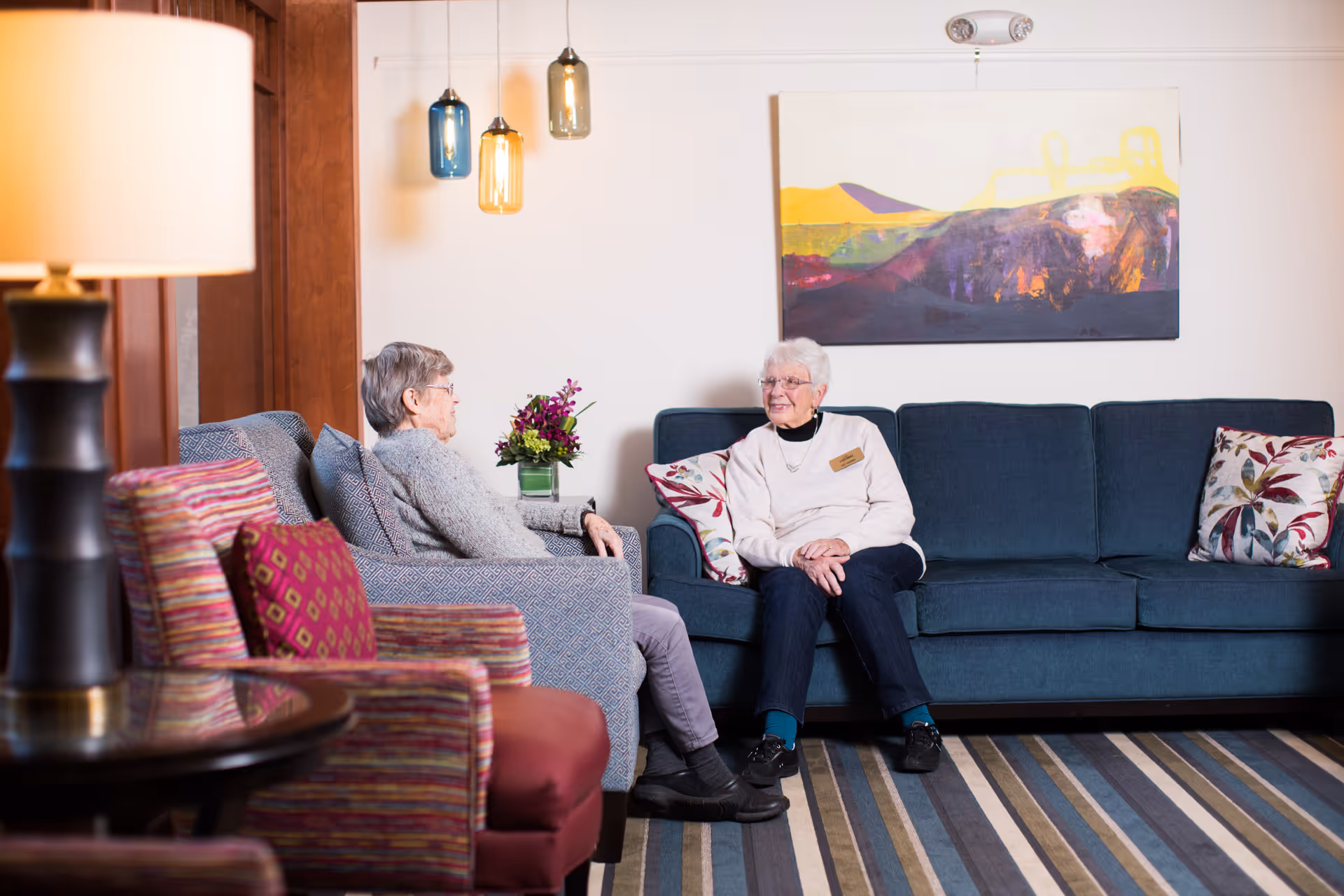 Two elderly women sitting and conversing in a cozy living room with a blue couch, patterned armchairs, a striped rug, a table lamp, hanging pendant lights, a vase with flowers, and a colorful abstract painting on the wall.