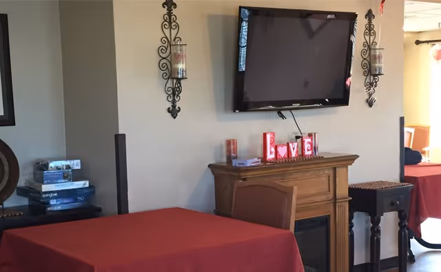 Small communal dining area with red-covered tables, a wall-mounted TV above a mantel displaying decorative 'LOVE' letters, and wall sconces.
