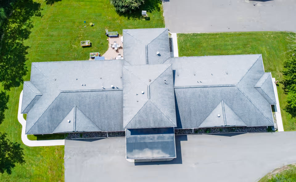 Aerial view of a single-story building with a gray roof, surrounded by green grass and trees, with paved driveways on two sides.