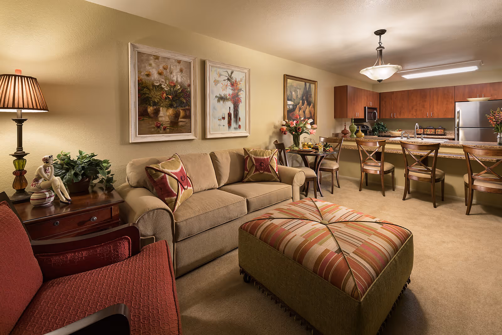 A cozy living room area in a senior living facility featuring a beige sofa with patterned cushions, a large striped ottoman, a red armchair, and a wooden side table with a lamp and decorative figurine. The room opens into a kitchen with wooden cabinets, a granite countertop with four bar stools, and a small dining table with two chairs. The walls are adorned with three framed floral paintings.
