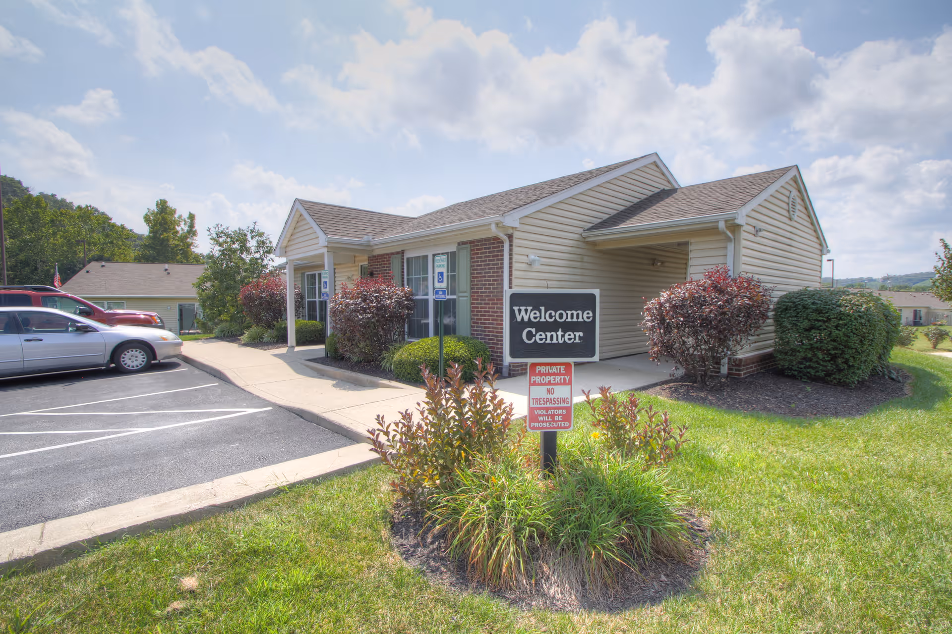Exterior view of a single-story building labeled as the Welcome Center with a parking lot and cars on the left side, surrounded by green grass, bushes, and a partly cloudy sky.