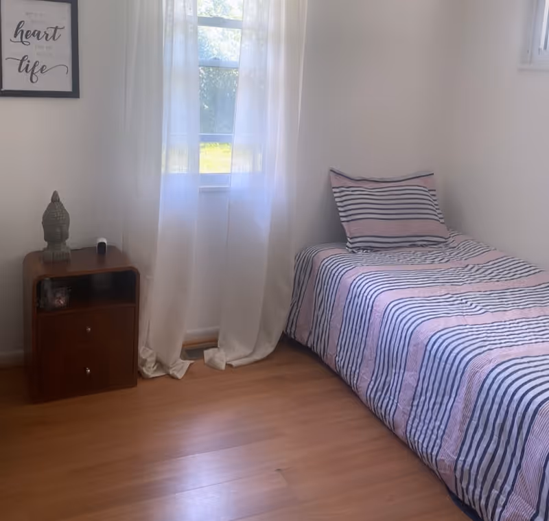 Cozy bedroom with striped bed, nightstand, Buddha statue, and sheer-curtained window.