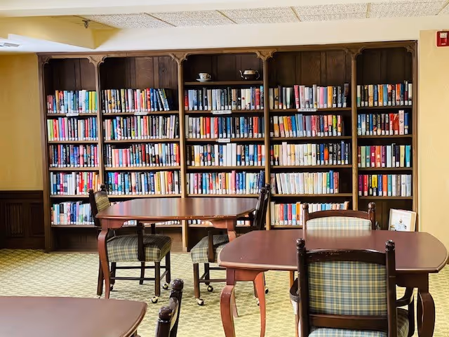 A cozy reading room with tall bookshelves along the back wall and wooden tables and chairs in the foreground.
