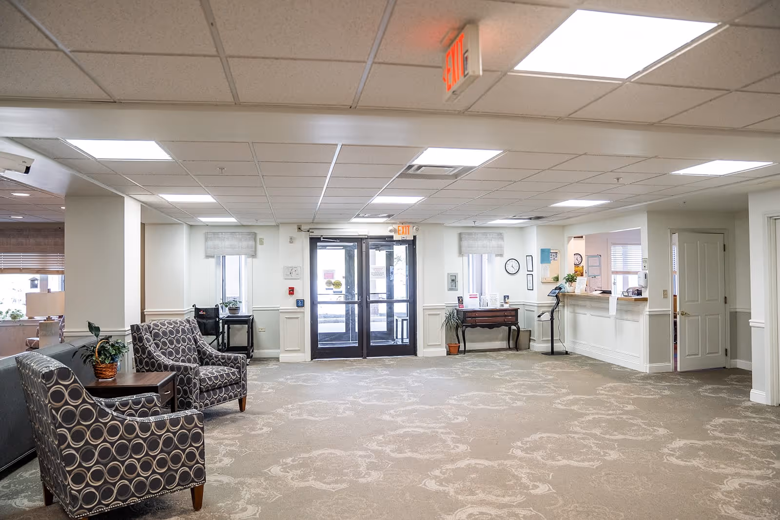 A spacious, well-lit lobby area with patterned carpet, two armchairs with circular designs, a small table with a plant, and a reception desk to the right. Double glass doors with exit signs above them are visible at the far end, along with windows and some wall clocks and framed items.