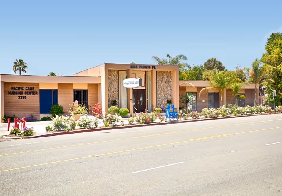 Exterior front view of Pacific Care Nursing Center building with a clear blue sky, palm trees, and landscaped bushes and flowers along the sidewalk.