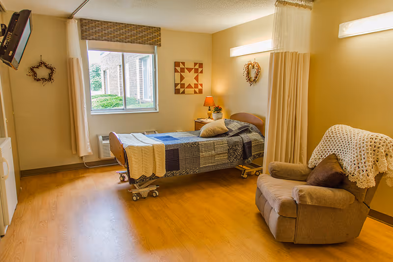 A well-lit room with a single hospital-style bed covered with a patchwork quilt and a pillow. Next to the bed is a small wooden nightstand with a lamp and a plant. A comfortable recliner chair with a crocheted throw and a cushion is positioned near the bed. The room has light-colored walls, a window with a view of greenery outside, and wooden flooring. Wall decorations include a star-shaped wreath and a geometric art piece. A curtain is partially drawn to separate the bed area.