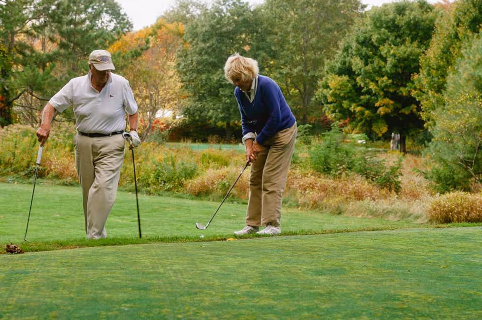 Two elderly people playing golf on a green golf course surrounded by trees and bushes with autumn foliage.