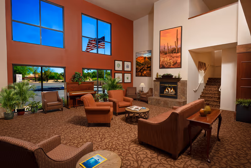 A spacious and warmly decorated living room area in an assisted living facility with large windows showing an American flag outside. The room features comfortable seating including armchairs and a sofa arranged around a coffee table, a fireplace with desert-themed artwork above it, a piano, several potted plants, and a carpeted floor. A staircase is visible in the background.