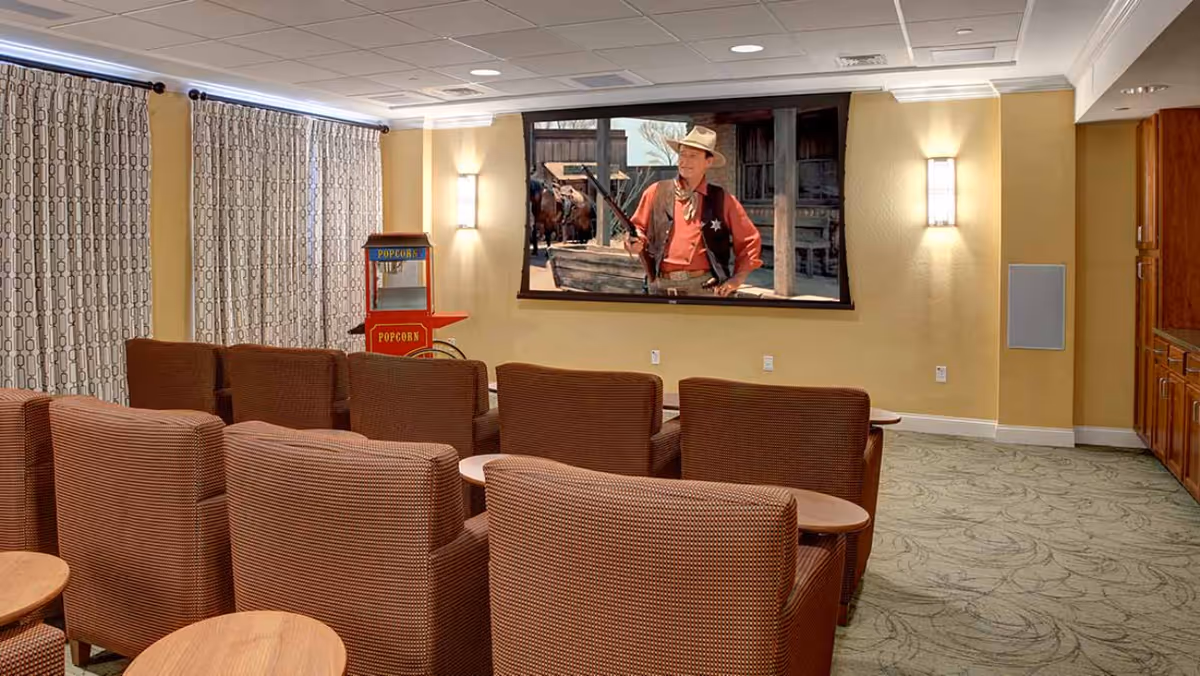 Small theater-style media room with rows of upholstered chairs facing a wall-mounted screen showing a western and a popcorn machine at the side.