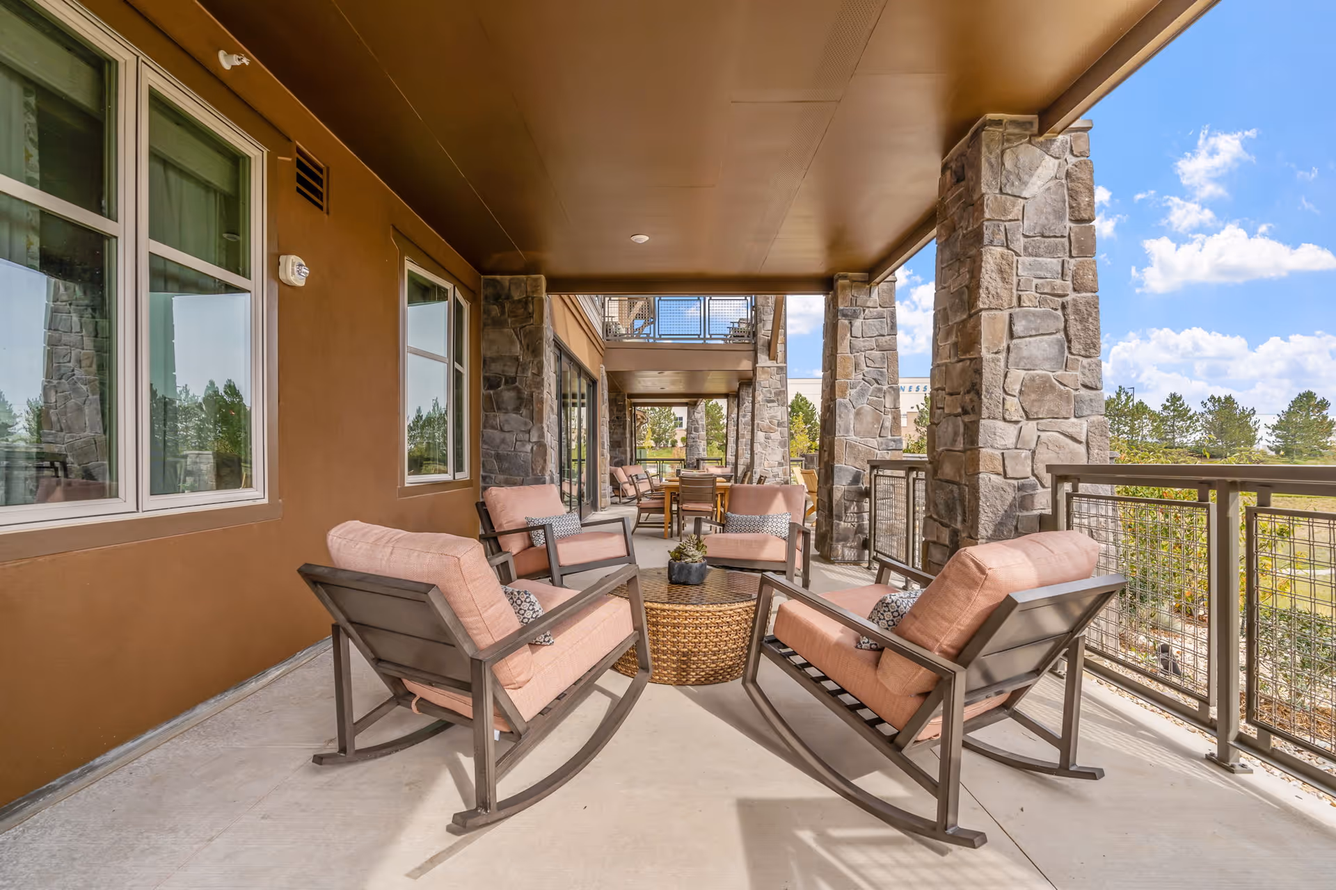 Covered outdoor patio with cushioned rocking chairs, a wicker table, stone pillars, and a view of the landscaped grounds.