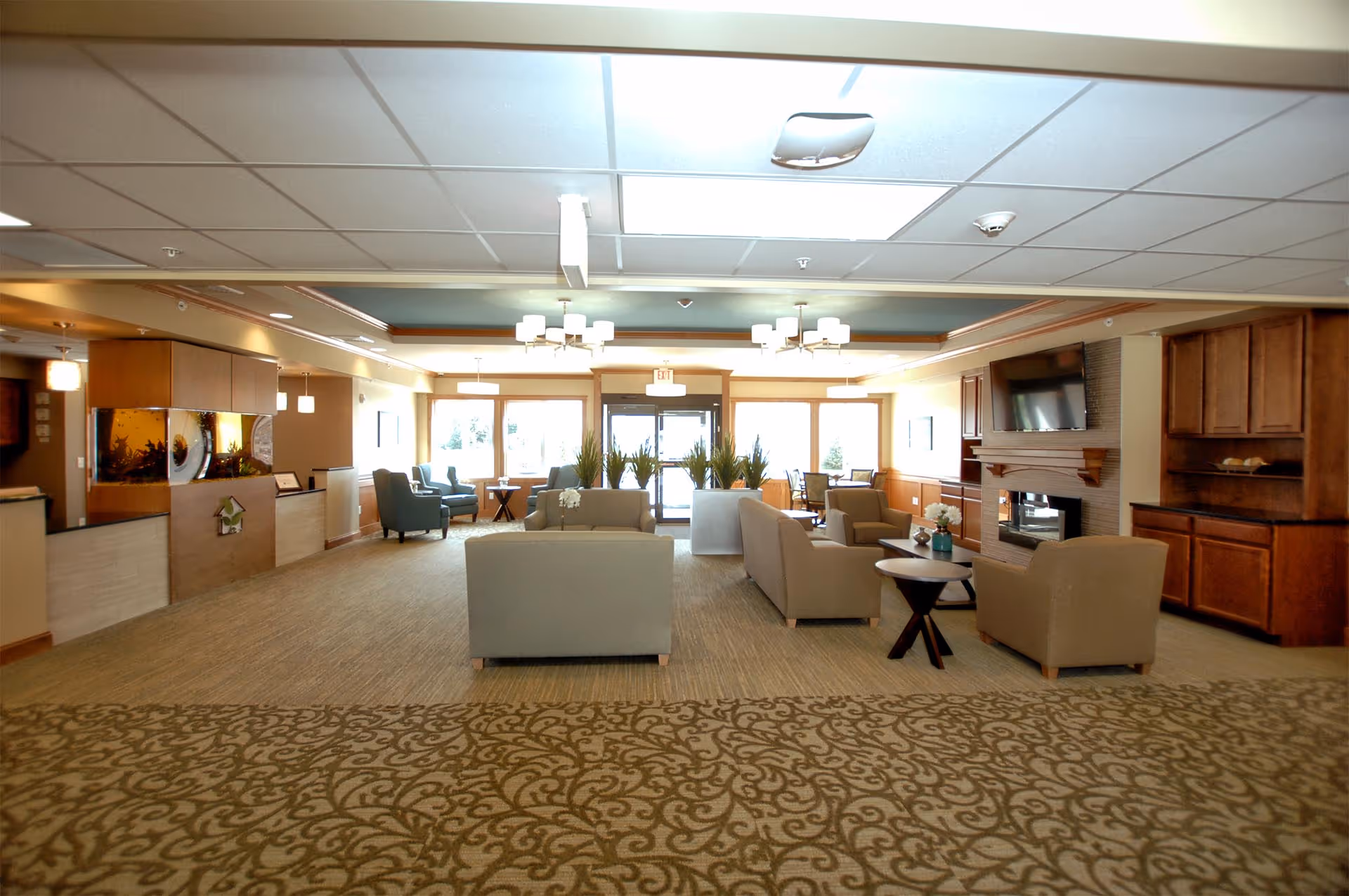 Spacious furnished common lounge with sofas and armchairs arranged around a fireplace and TV, a reception desk to the left, and large windows at the far end.
