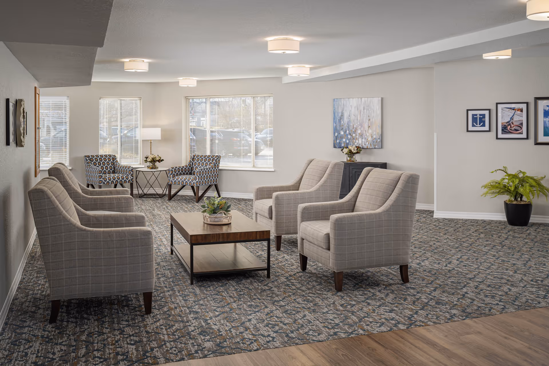 A bright and spacious senior living facility lounge area with six beige upholstered armchairs arranged around a wooden coffee table. The room has large windows letting in natural light, patterned carpet flooring, and light-colored walls adorned with framed artwork. There is a small table with a lamp and flowers near the windows, and a potted plant in the corner.