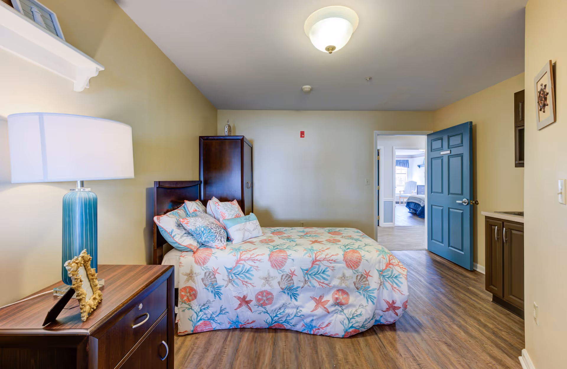 A bedroom in a senior living facility with a bed covered in a colorful seashell and coral patterned bedspread. There are multiple pillows on the bed. To the left is a wooden nightstand with a blue lamp and a decorative picture frame. The floor is wood, and the walls are painted light yellow. A blue door is open, showing another room with a bed and window in the background.