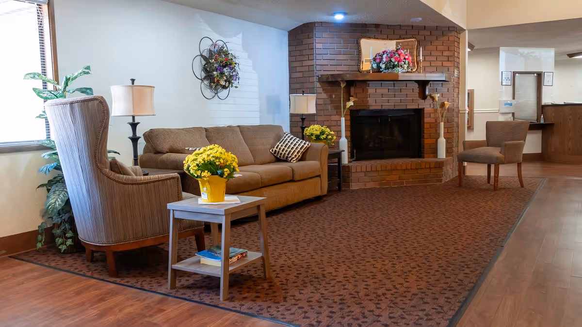 Cozy senior living lounge with a sofa, armchairs, small table with flowers, and a brick fireplace.