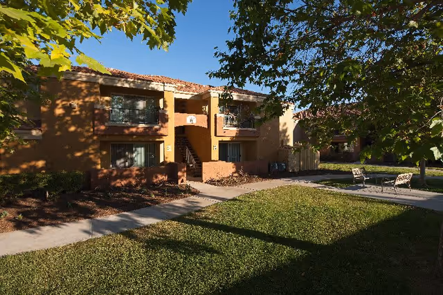 Two-story stucco apartment building with balconies facing a grassy courtyard, paved walkway, and patio chairs beneath leafy trees.