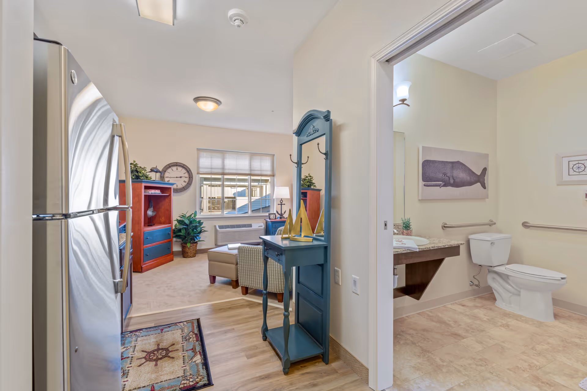 View of a senior living facility apartment showing a kitchen area with a stainless steel refrigerator on the left, a small blue hall table with decorative sailboats and a mirror, leading into a living room with a window, armchair, ottoman, and wooden furniture. To the right, there is an open doorway revealing a bathroom with a toilet, sink with granite countertop, grab bars, and wall art featuring a whale and a compass.