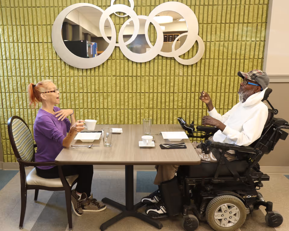 Two elderly individuals sitting across from each other at a dining table in a room with a green textured wall and a decorative circular mirror. One person is seated in a wheelchair wearing a white hoodie and cap, while the other person is wearing a purple top and glasses, holding a cup.