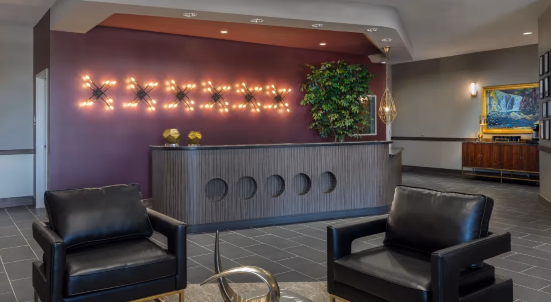 Modern reception area with a wooden front desk featuring circular cutouts, a purple accent wall with decorative lighting, two black leather armchairs, a large green plant, and a sideboard with a framed painting of a waterfall.
