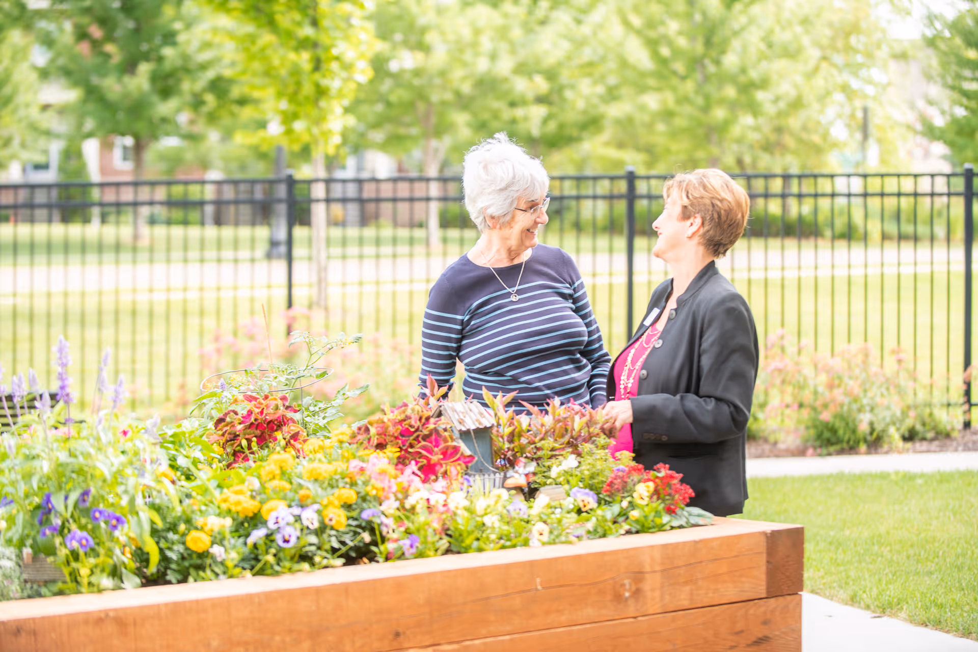 Two women smiling and talking to each other while standing next to a raised wooden garden bed filled with colorful flowers and plants, with a black metal fence and green trees in the background.