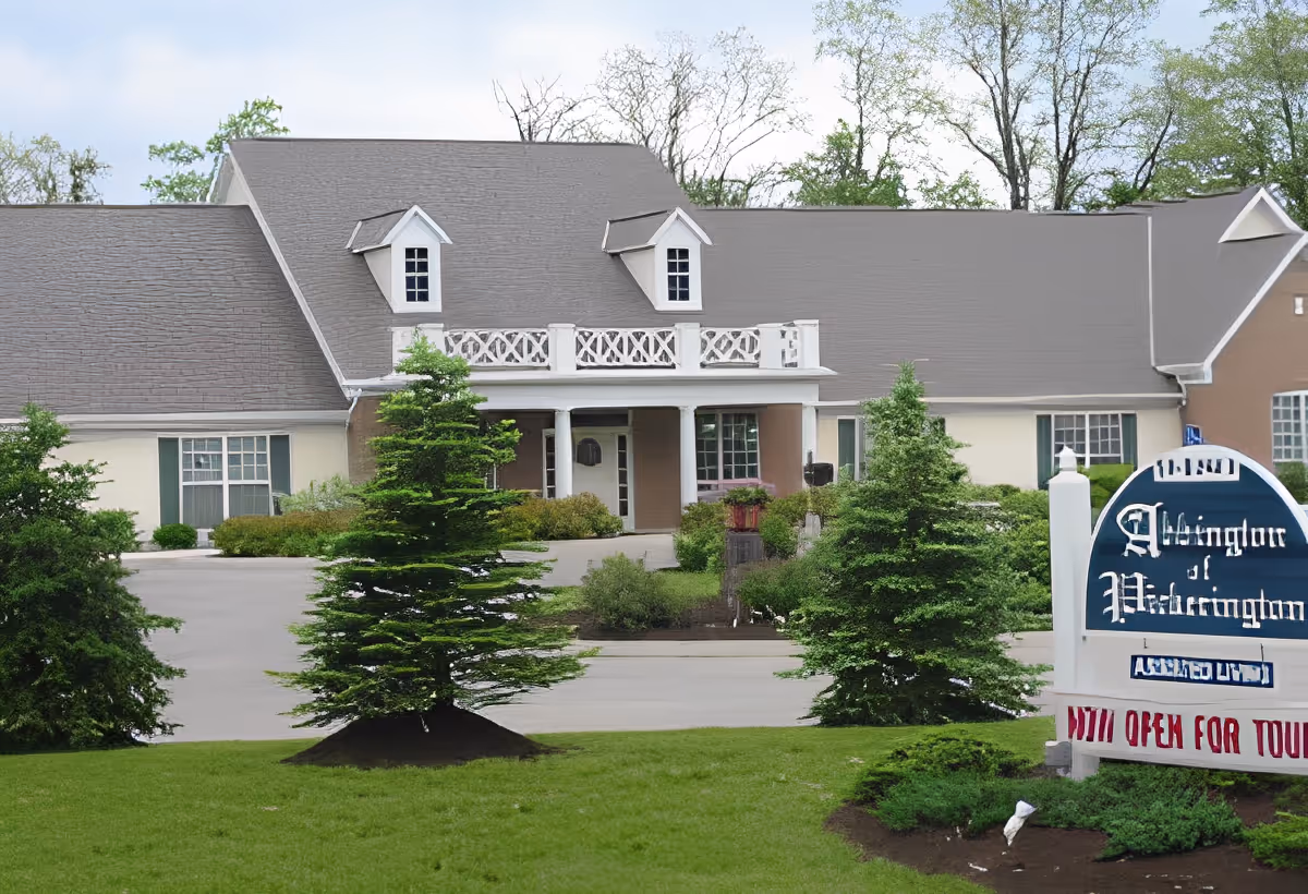 Exterior view of Abbington of Pickerington, a senior living facility with a large building featuring a gray roof, white columns at the entrance, and surrounded by green trees and shrubs. A sign in front displays the facility's name and indicates it is open for tours.