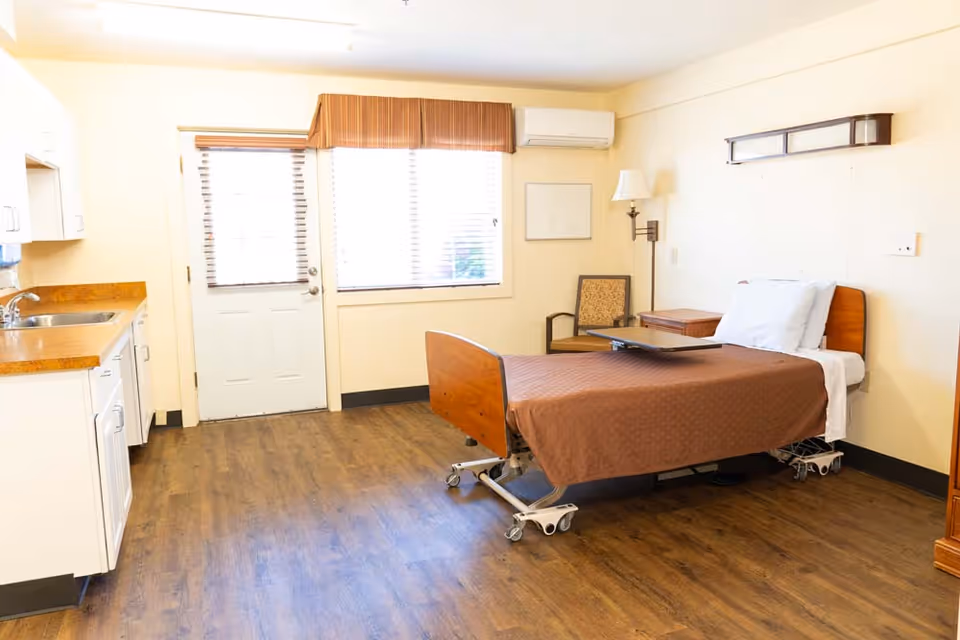 A bright room with a single hospital-style bed covered with a brown blanket and white pillow. The room has wooden flooring, a small kitchenette with a sink and cabinets on the left, a door and window with blinds and a brown valance, a chair, a small table, and a wall-mounted lamp.