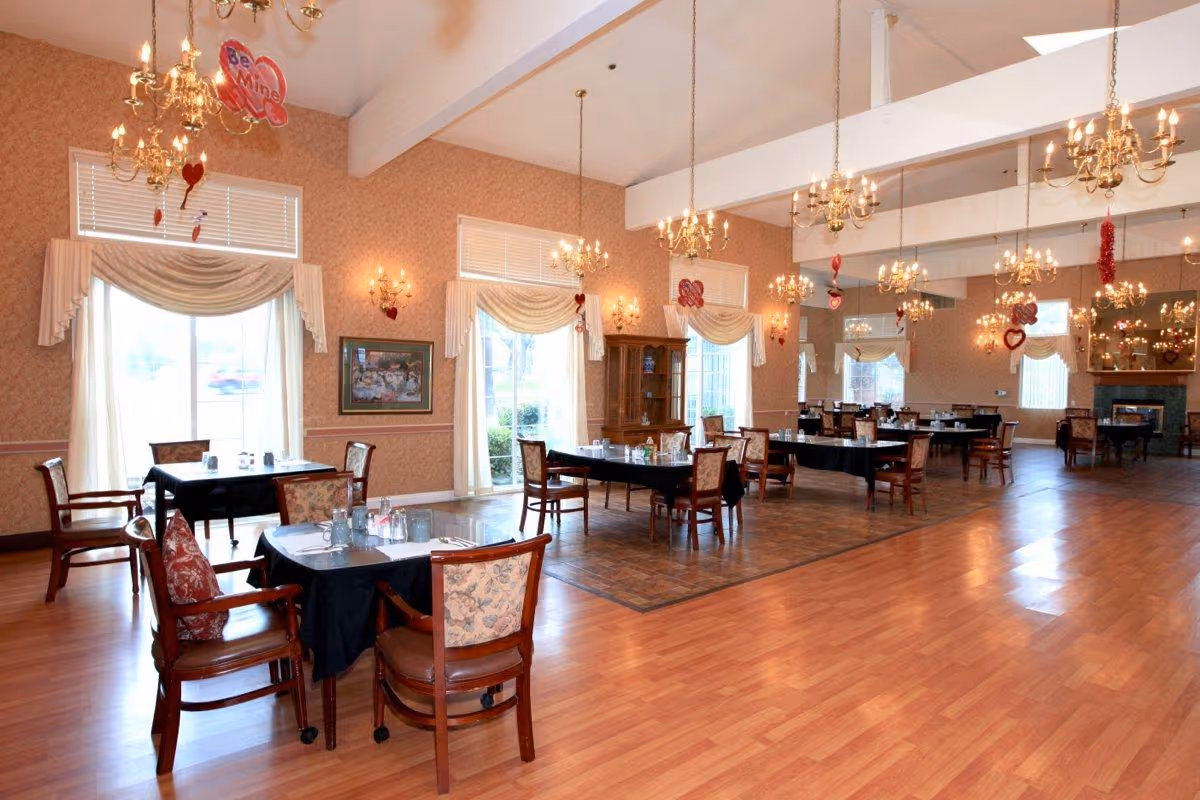 Spacious dining room with wooden floors and several tables covered with black tablecloths, each surrounded by wooden chairs with patterned cushions. The room is decorated with chandeliers and wall sconces, and has large windows with draped curtains letting in natural light. Valentine's Day decorations, including heart-shaped balloons and hanging ornaments, are visible throughout the room.