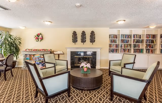A cozy senior living common area with four light green cushioned armchairs arranged around a round dark coffee table with a pink flower centerpiece. Behind the seating area is a fireplace with three decorative black leaf wall hangings above it. To the right, there is a built-in bookshelf filled with books. To the left, there is a table with board games and a large potted plant next to it. The room has a patterned carpet and soft lighting from ceiling fixtures.