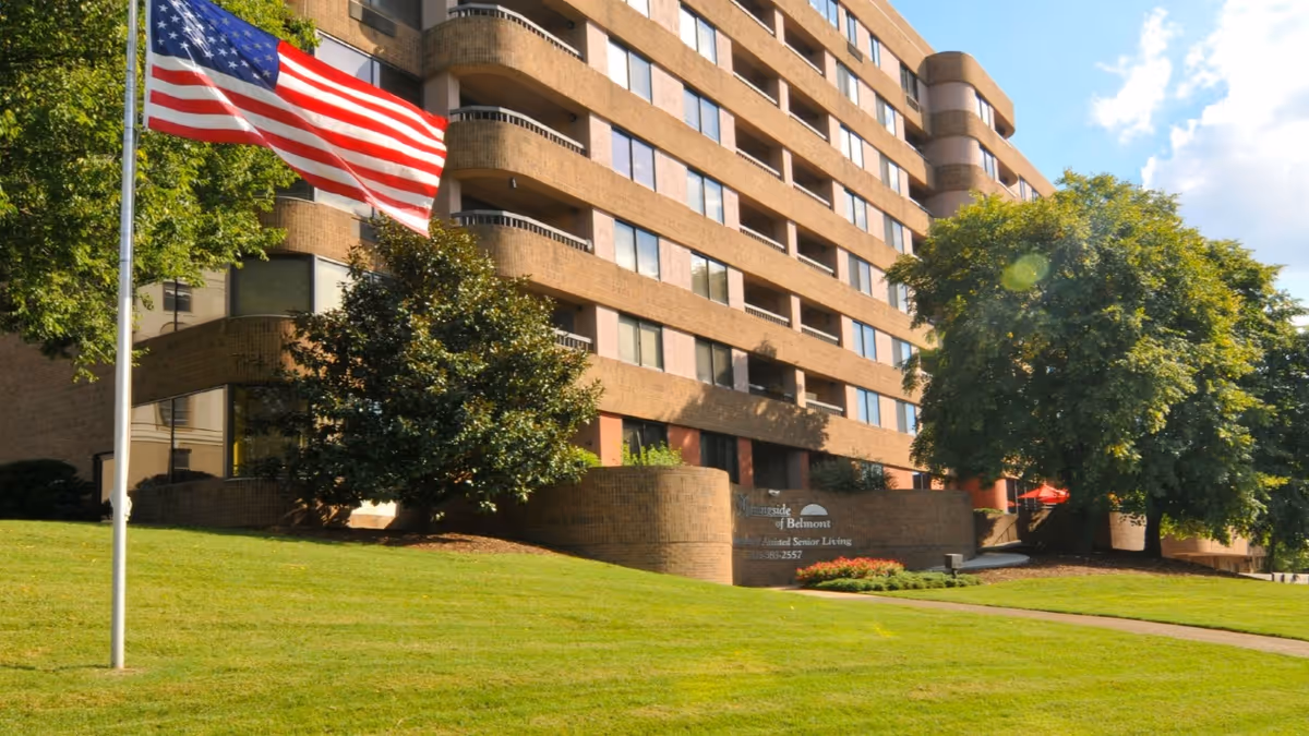 Exterior view of a multi-story brick building with large windows, surrounded by green grass and trees. An American flag is flying on a flagpole in the foreground. A sign near the entrance reads 'Morningside of Belmont Assisted Senior Living.'