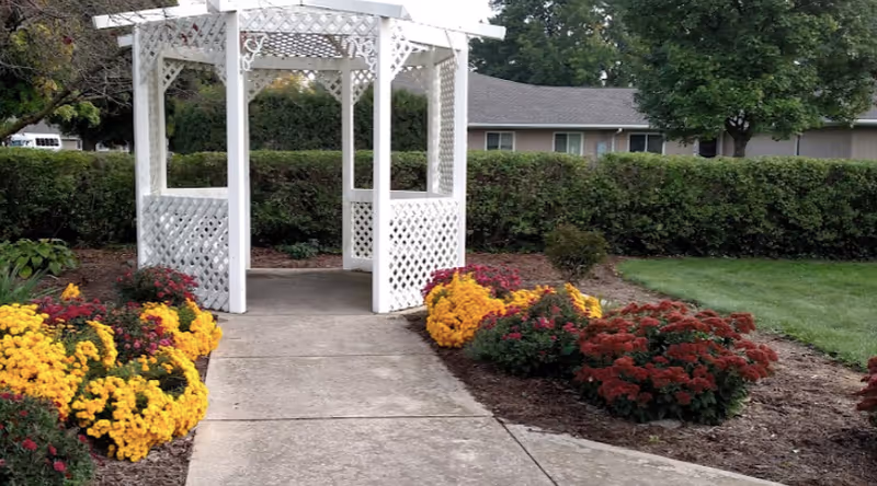 A white wooden gazebo with lattice sides situated at the end of a concrete pathway, surrounded by colorful flower beds with yellow, red, and pink flowers, green bushes, and a grassy lawn. In the background, there is a hedge and a single-story building partially visible.