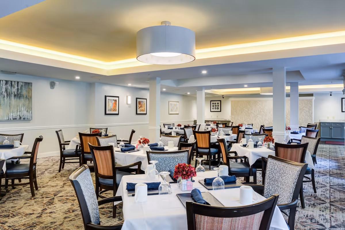 A spacious dining room with multiple tables covered in white tablecloths, each set with napkins, glasses, and mugs. The room features patterned carpet, modern light fixtures, and framed artwork on the walls, creating a welcoming atmosphere.
