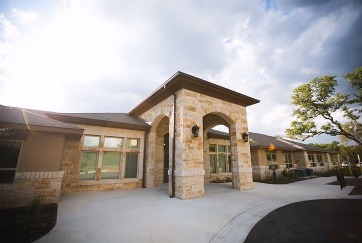 Exterior view of a single-story building with stone and beige walls, large windows, and an arched entrance. The sky is partly cloudy with sunlight shining through, and there is a tree on the right side near the driveway.