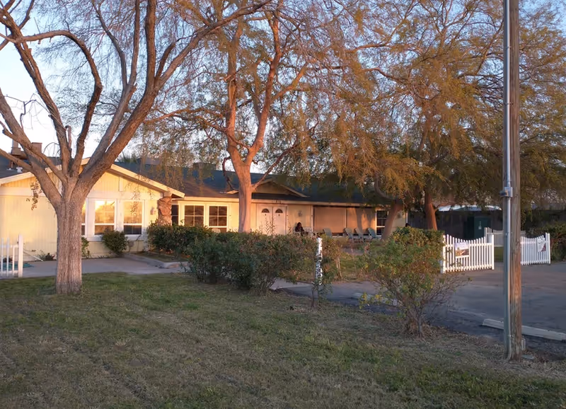 Single-story assisted living facility exterior with trees, front yard, and white picket fence.