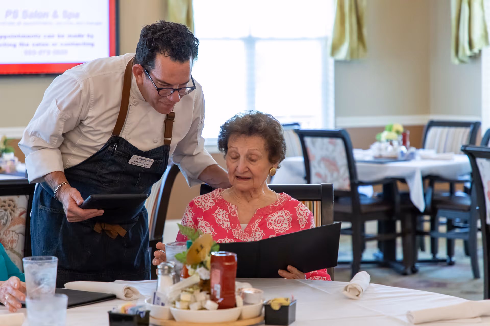 A server leans over to help an elderly woman seated at a dining table as she looks at a menu in a bright dining room.