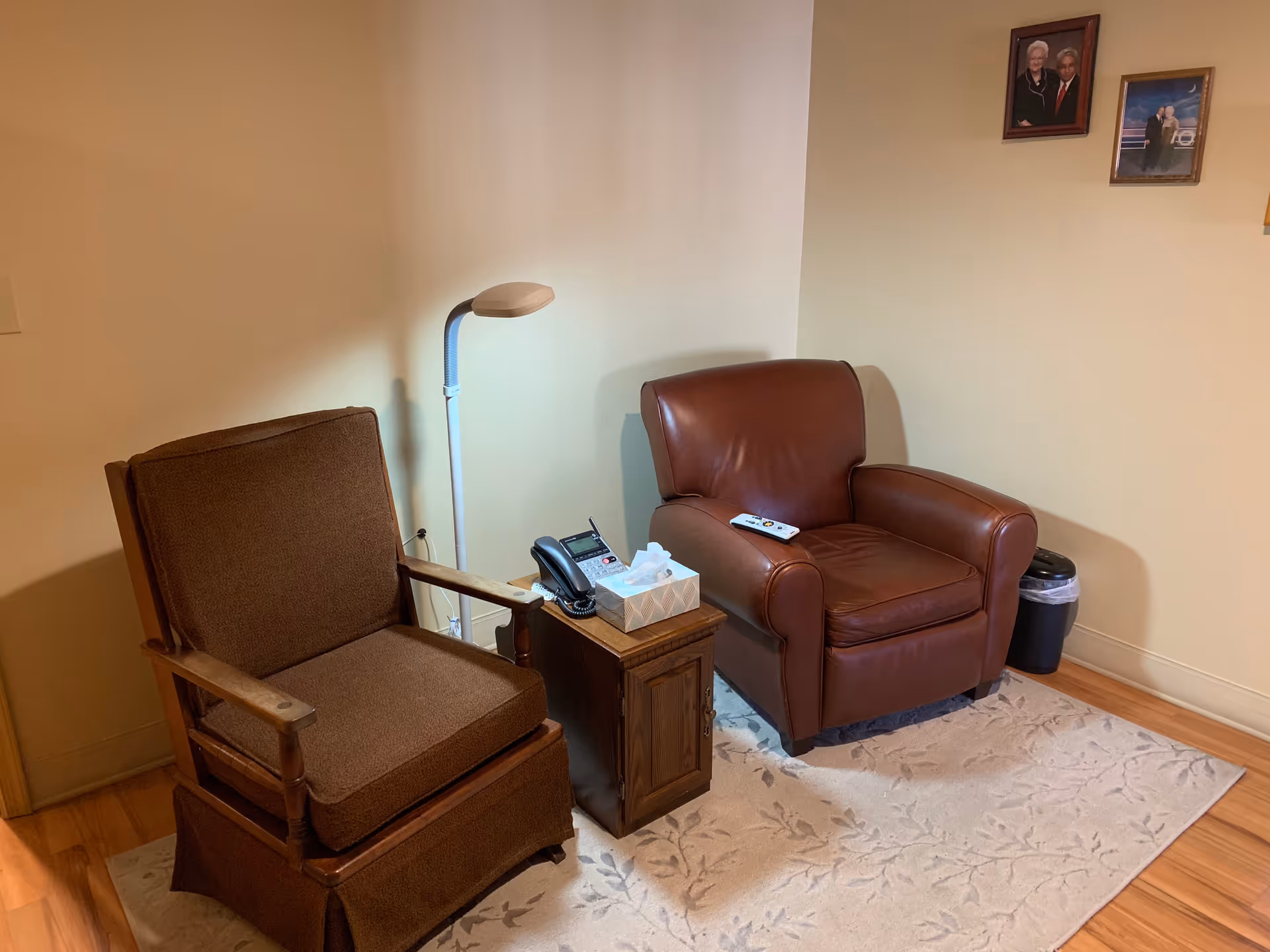 A cozy corner in a living area with a brown cushioned wooden armchair and a brown leather armchair placed on a light-colored patterned rug. Between the chairs is a small wooden side table holding a telephone and a box of tissues. A floor lamp stands behind the side table. On the wall above the leather chair are two framed photographs. A small trash bin is placed near the wall.