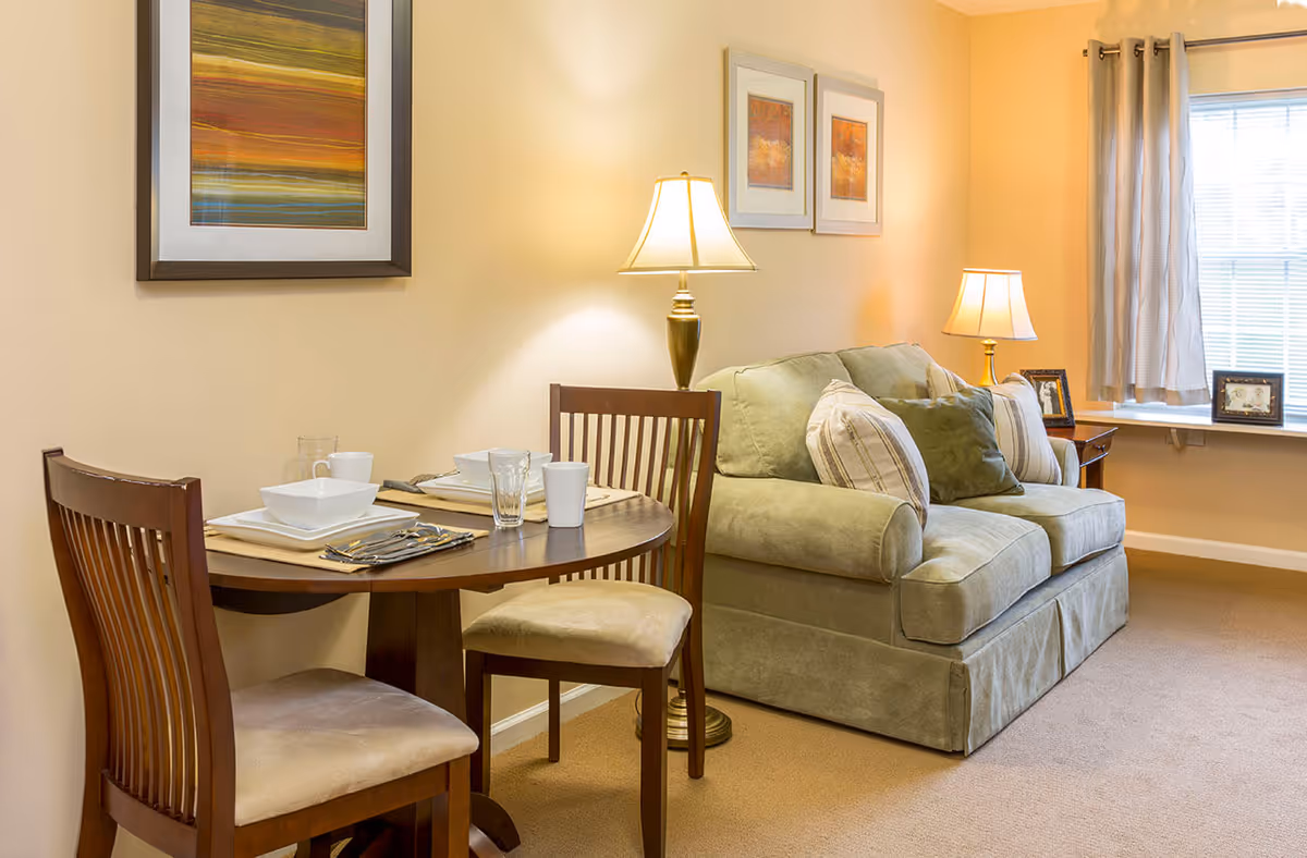 Cozy living room with a small round dining table set, a green loveseat, lamps, and framed wall art.