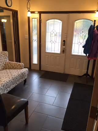 A home's entry foyer with double decorative glass front doors, tiled floor, patterned armchair, coat rack with jackets, and pendant lights.