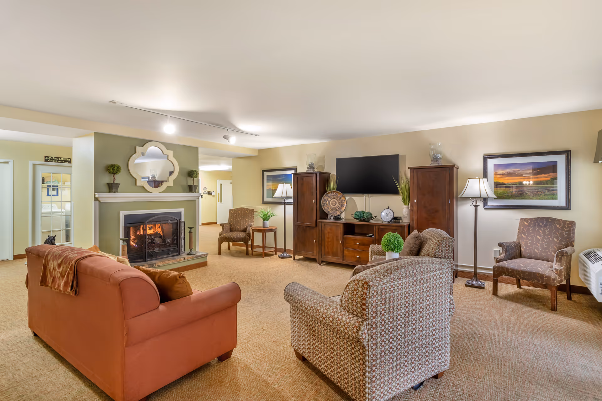 A cozy living room with a lit fireplace on the left wall, a large flat-screen TV mounted on a wooden cabinet, and several upholstered chairs and a sofa arranged around the room. The walls are painted light yellow and green, with framed artwork and decorative plants. The room is well-lit with ceiling lights and a floor lamp.