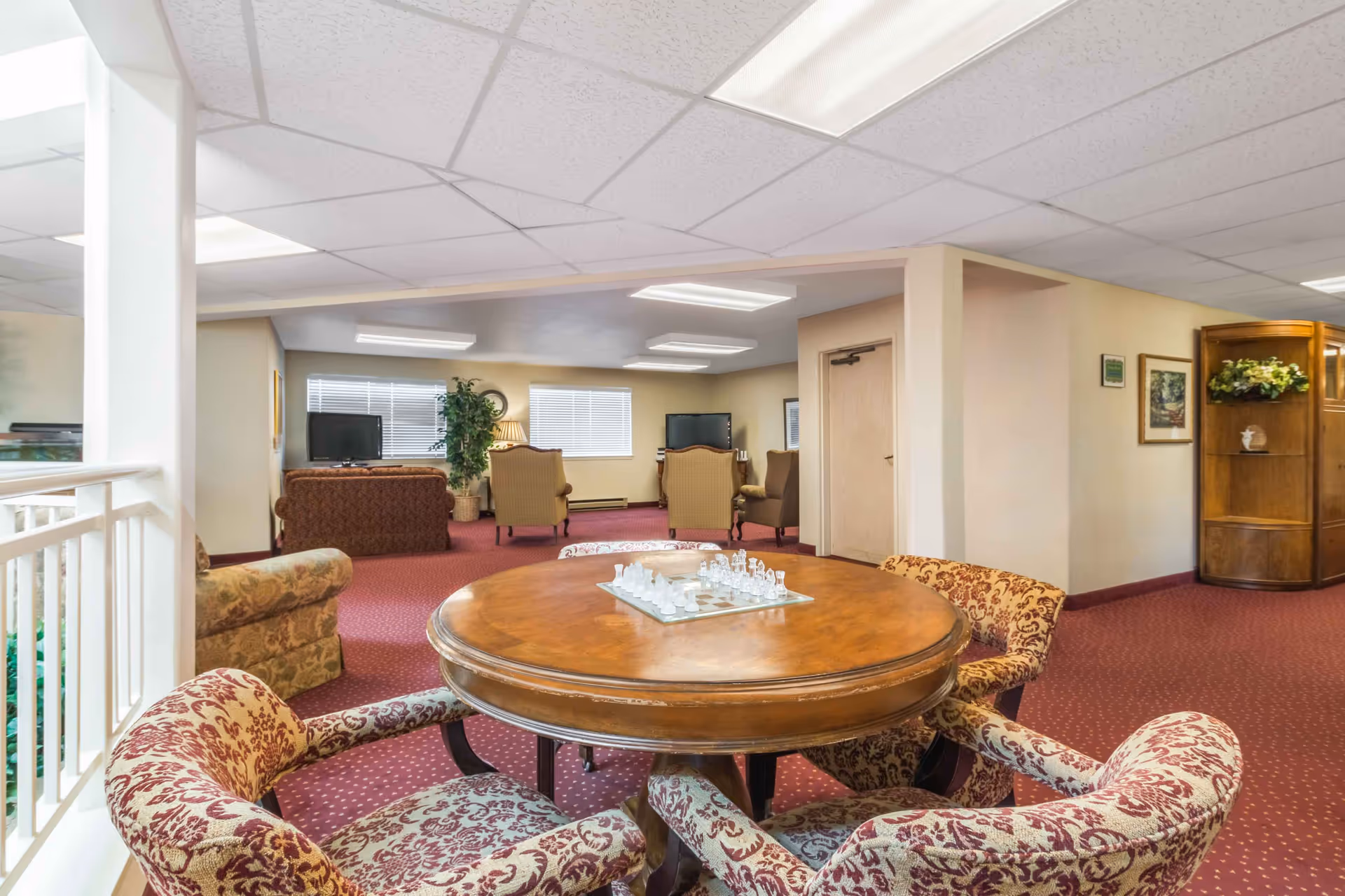 Carpeted senior living common room with a round wooden game table and upholstered chairs in the foreground and a seating area with TVs in the background.