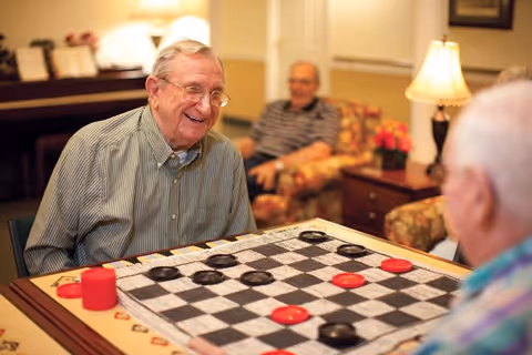 Two elderly men playing a game of checkers at a table in a cozy common area with warm lighting, floral armchairs, a lamp, and a piano in the background.
