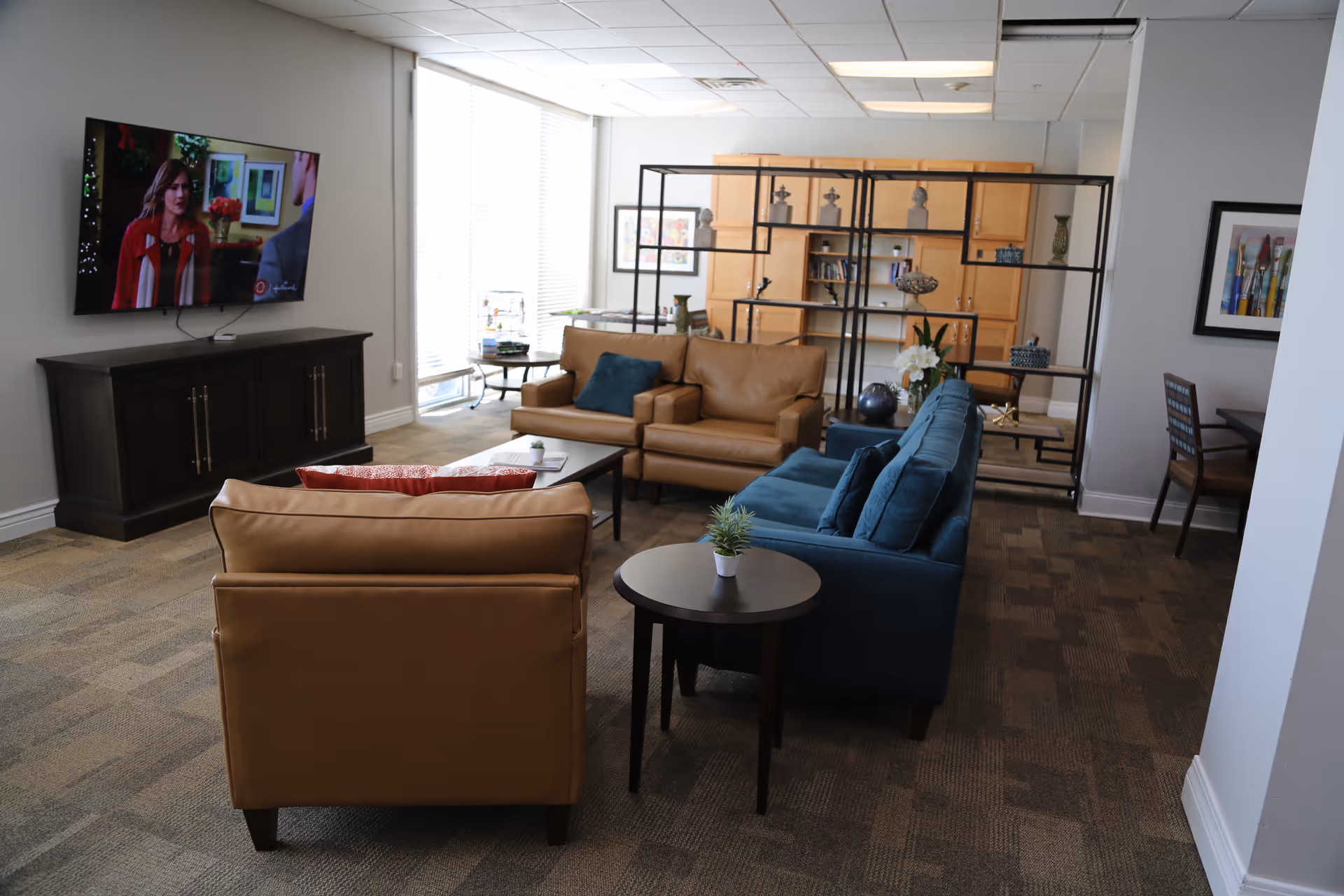 A cozy living room area with a tan leather armchair and loveseat, a blue fabric sofa, and a dark wood coffee table. A flat-screen TV is mounted on the wall showing a scene from a show. Behind the seating area is a black metal and wood shelving unit with decorative items and a wooden cabinet. Large windows with blinds allow natural light to fill the room. The carpet is patterned in neutral tones, and there is a small round side table with a potted plant in the foreground.