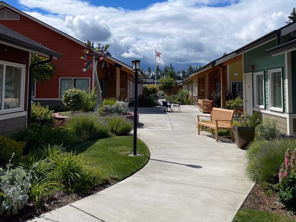 Sunny courtyard walkway between single-story colorful cottages with benches, landscaping, and outdoor seating under a partly cloudy sky.