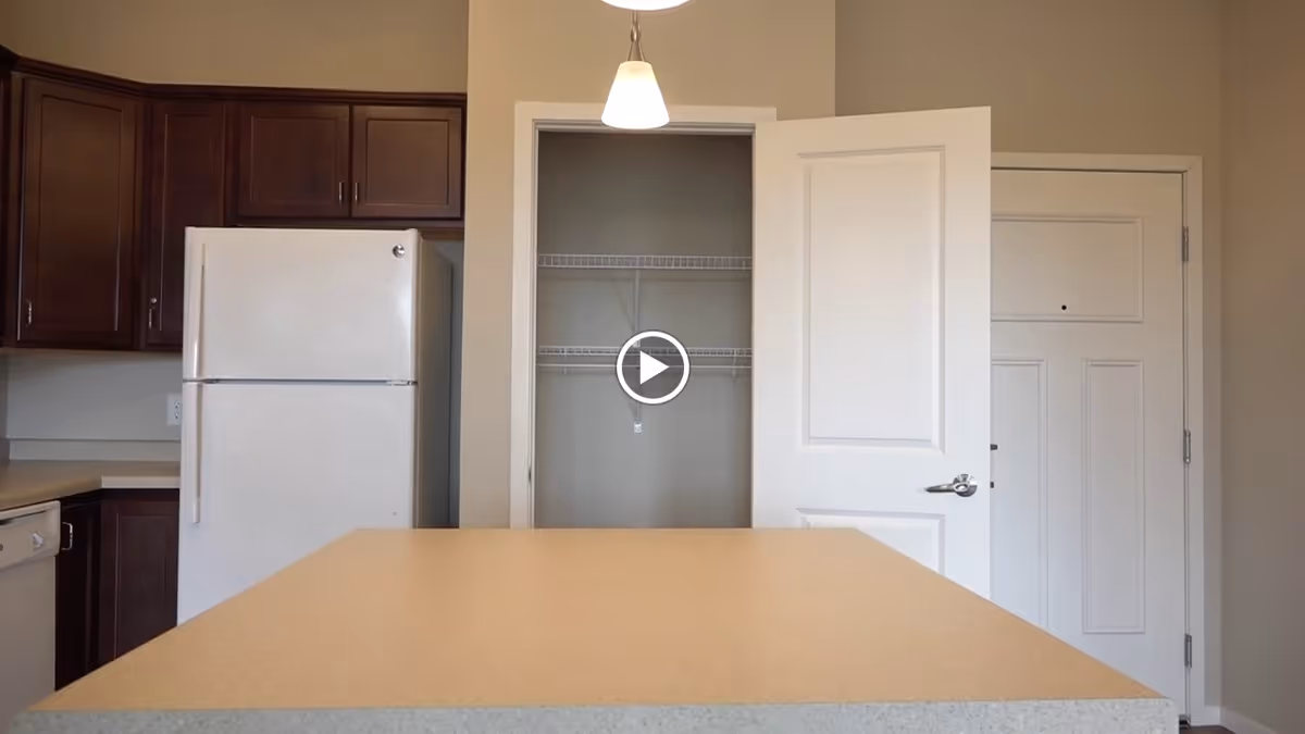 Interior view of a kitchen area featuring a beige countertop island in the foreground, dark wood cabinets, a white refrigerator, a pantry with an open door showing wire shelving, and a closed white door next to the pantry. Two pendant lights hang from the ceiling.