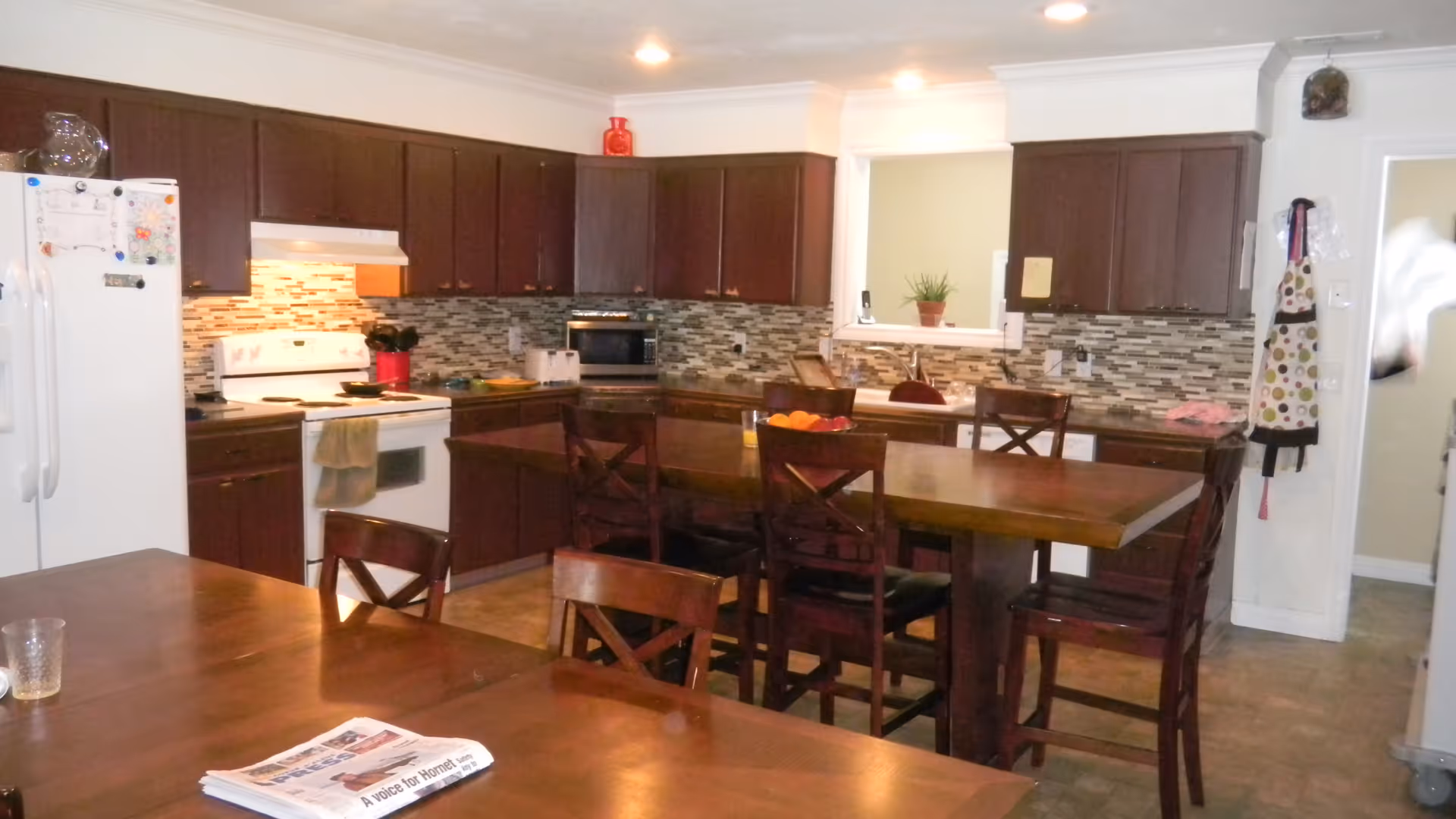 Open kitchen with dark wood cabinets, a central wooden island surrounded by chairs, a dining table in the foreground, white appliances, and a tiled backsplash.
