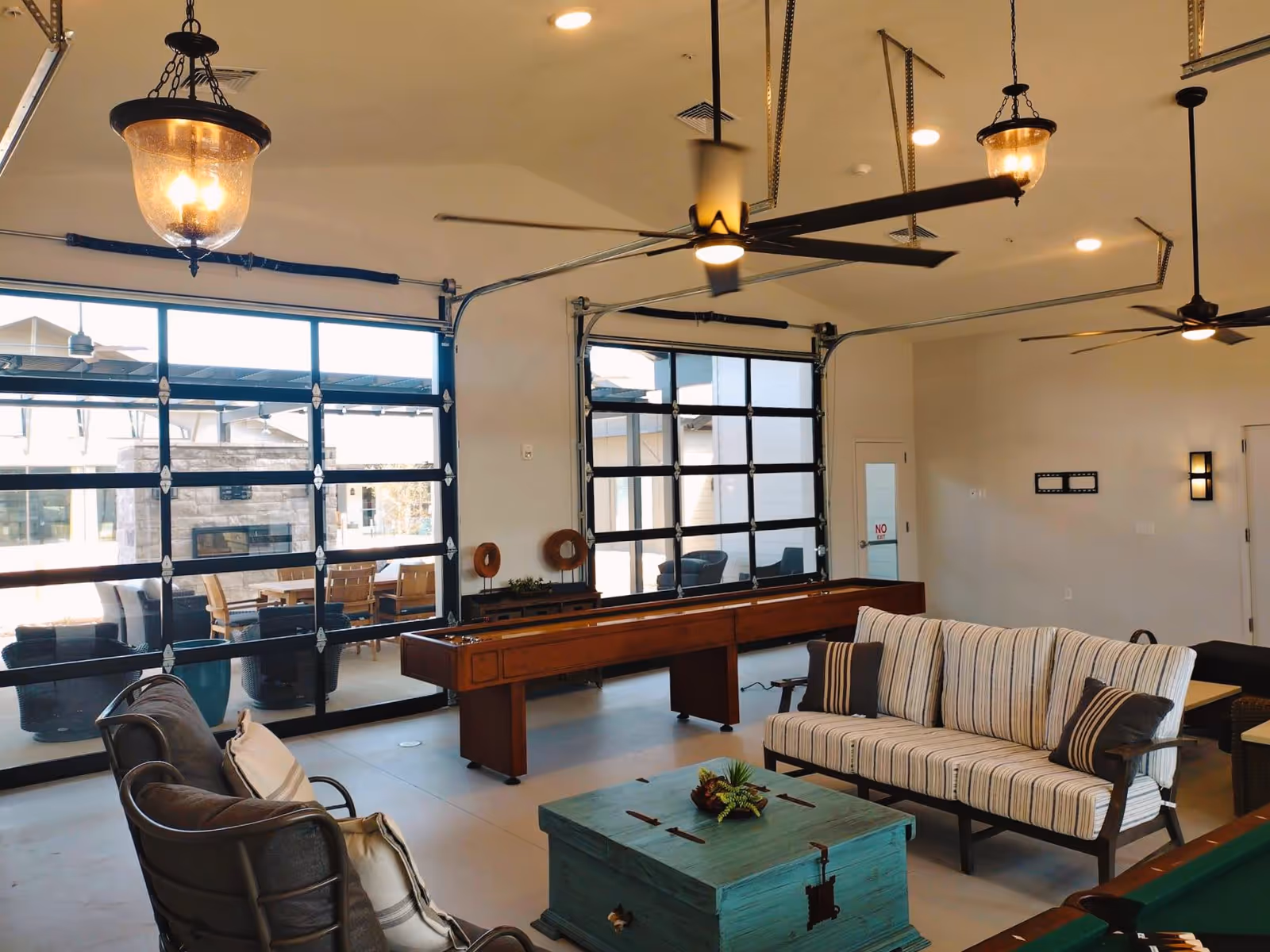 A spacious common area with large glass garage-style doors opening to an outdoor patio. Inside, there are two ceiling fans, a shuffleboard table, a striped cushioned sofa, a cushioned armchair, and a turquoise wooden chest used as a coffee table. The outdoor patio has a dining table with chairs and a stone fireplace.