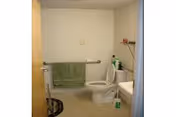 A small bathroom with a white toilet, a green towel hanging on a metal bar, a white sink with cleaning supplies on the counter, and a beige door partially open.