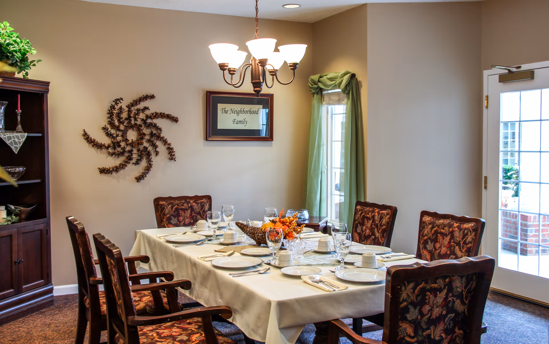 A dining room with a rectangular table set for six people. The table is covered with a white tablecloth and has plates, glasses, napkins, and a centerpiece with autumn-themed decorations. There are six wooden chairs with floral upholstery around the table. On the wall, there is a framed sign that reads 'The Neighborhood Family' and a decorative wall art piece. A chandelier with five lights hangs above the table. A window with green curtains and a glass door letting in natural light are visible in the background.