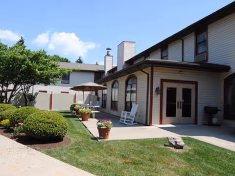 Exterior courtyard and entrance of a two-story senior living building with patio furniture, rocking chair, potted plants, and landscaped shrubs.